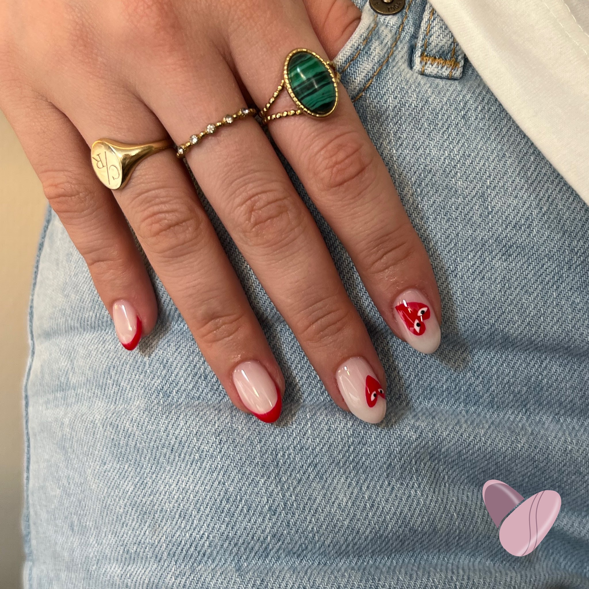 Nude nagels met rode French en rode hartjes, hand op een spijkerbroek met gouden ringen
